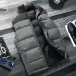 Una giacca trapuntata grigia e nera, senza maniche e senza busto, con tasche semi-aperte con cerniera, stesa su un pavimento a strisce grigio scuro. Ai lati, un berretto nero, una scarpa da ginnastica nera e un po' di targa.