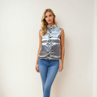 Donna bionda in piedi contro una parete beige che indossa jeans skinny blu e un piumino argentato senza maniche.