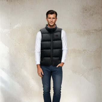 Uomo dai capelli scuri che indossa jeans blu scuro, top bianco a maniche lunghe e giacca nera trapuntata senza maniche. In piedi, con le spalle rivolte a una parete beige.