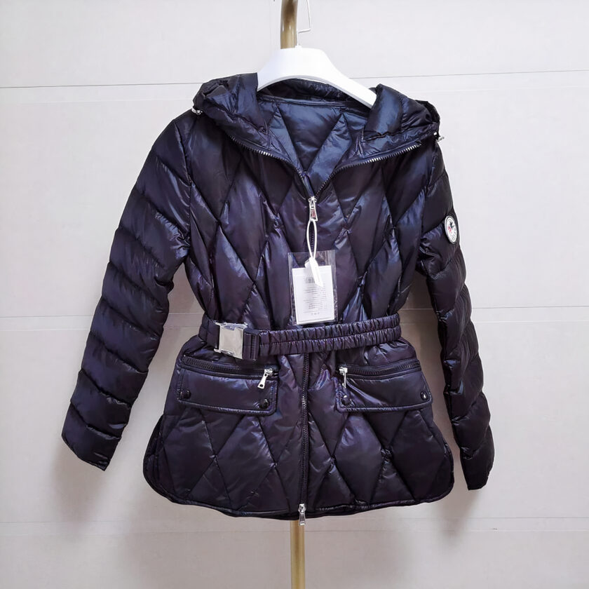 Vediamo un piumino nero trapuntato e con cintura, dalla vestibilità slim e con cappuccio. È esposto su una cintura appesa, su uno sfondo grigio. L'etichetta è attaccata alla cerniera.