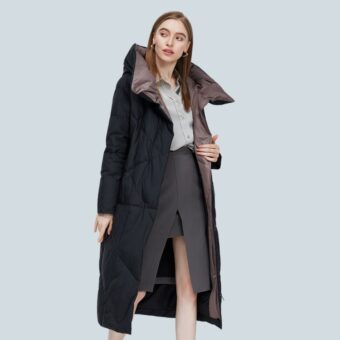 Donna con capelli lunghi e lisci color castagna che indossa un lungo piumino nero aperto con un grande colletto marrone all'interno. Sotto indossa una camicia grigio chiaro e una gonna a tubino grigio scuro.