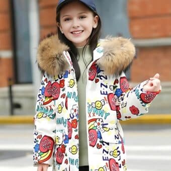 Giovane ragazza in una strada che indossa un piumino bianco con cappuccio per adolescenti Il piumino ha un cappuccio di pelliccia beige Motivi colorati sono stampati su tutto il cappotto La giovane ragazza indossa un berretto blu navy.