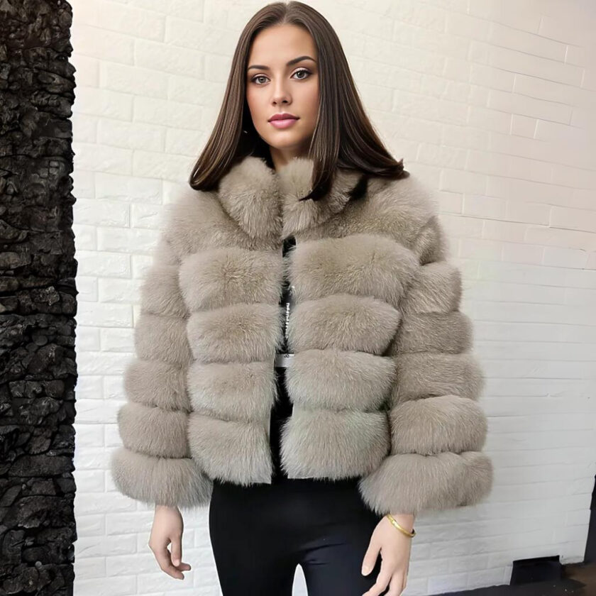 Donna dai capelli castani che dà le spalle a un muro di pietra bianca, indossa un piumino di pelliccia sintetica beige e pantaloni neri.