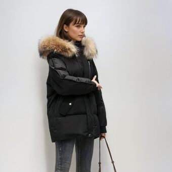 Donna dai capelli castani con frangia, in piedi davanti a una parete bianca/grigia, indossa un piumino nero con cappuccio e pelliccia marrone trapuntata a metà lunghezza. Con la mano sinistra tiene la tracolla di una borsetta.