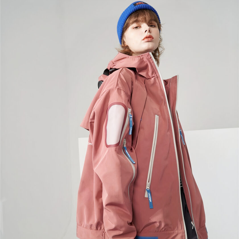 Donna con cappello blu che indossa una giacca da sci rosa di media lunghezza per donna su sfondo grigio.