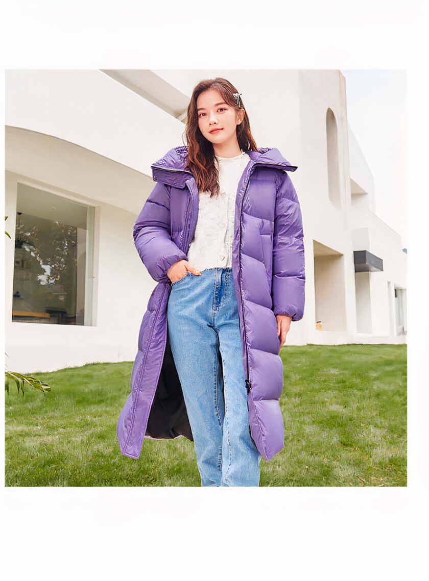 Giovane donna bruna dai capelli lunghi che indossa un maglione bianco sotto una lunga e spessa giacca da donna viola aperta con cappuccio, con una mano nella tasca dei jeans blu. Sullo sfondo c'è del verde e un edificio bianco con un bovindo.