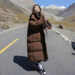 Donna che indossa un lungo piumino marrone. È in piedi su una strada in mezzo alle montagne.
