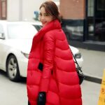Foto di una donna che indossa un lungo piumino rosso e cammina per strada, con un'auto bianca dietro a sinistra e un dissuasore giallo a destra.