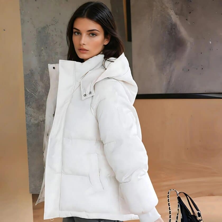 Donna dai capelli castani di profilo che indossa un piumino bianco corto con cappuccio. Una borsa dietro di lei e una parete beige e grigia.