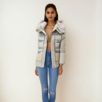 Donna con i capelli scuri che indossa jeans blu, un crop top marrone e un piumino beige di media lunghezza con cappuccio e tasche laterali. È in piedi e dà le spalle a una parete beige.