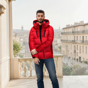 Uomo dai capelli scuri in piedi su una terrazza in città, indossa jeans blu e una giacca rossa imbottita con cappuccio. Sullo sfondo un edificio in stile parigino.