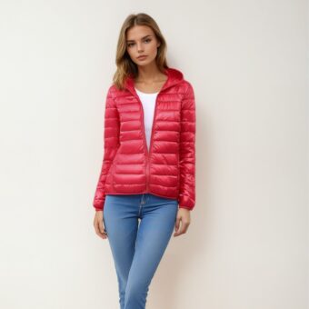 Donna bionda in piedi con le spalle al muro beige, indossa jeans skinny blu, un top bianco e, sopra, un leggero e caldo piumino rosso, chiuso con la zip a un terzo, che lascia intravedere il top bianco.