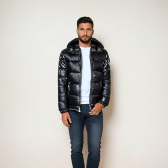 Un uomo dai capelli scuri non rasati che indossa jeans blu, una maglietta bianca e una giacca nera lucida con cappuccio trapuntato. È in piedi davanti a un muro beige.