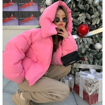 Donna accovacciata con occhiali da sole davanti a un albero di Natale con piumino rosa e cappuccio in testa.