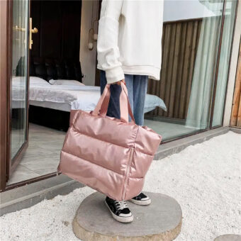 Grande borsa trapuntata di colore rosa metallizzato portata da una persona. La persona, che indossa una felpa bianca e jeans blu, si trova all'esterno su un tronco di legno sottile su un pavimento di ghiaia bianca. Si intravede una camera da letto con un letto sullo sfondo dietro una finestra a golfo.