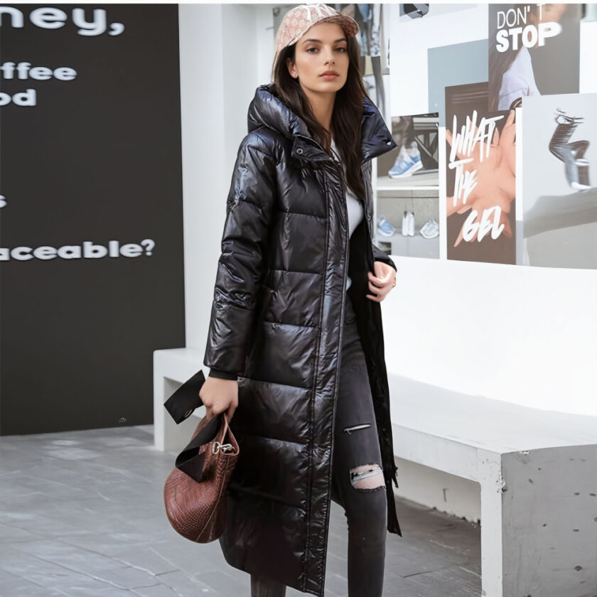 Donna in piedi con un muro con scritte sullo sfondo a destra e un muro nero a sinistra con scritte bianche. Una panchina di pietra grigia dietro la donna. La donna indossa pantaloni neri con un buco sul ginocchio sinistro, un berretto beige, un lungo piumino nero con cappuccio e una borsa nella mano destra.