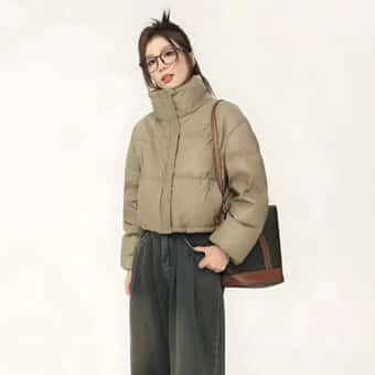 Donna asiatica con i capelli legati all'indietro e con gli occhiali in piedi. Indossa pantaloni neri e una giacca corta in similpelle color cachi. Le mani sono nelle tasche dei pantaloni. Sulla spalla sinistra porta una borsa nera e marrone. Su sfondo beige.
