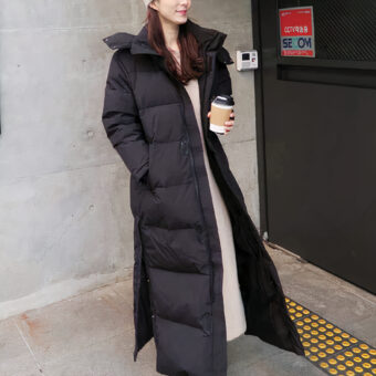 Donna dai capelli castani in una tromba delle scale, davanti a una porta di emergenza, con indosso un grande piumino nero, caldo e con cappuccio.