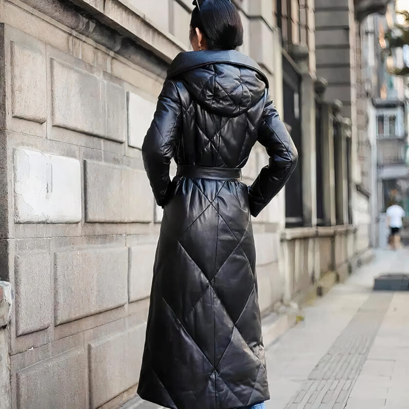 Una donna dai capelli scuri è vista da dietro in strada. Porta uno chignon. Indossa una lunga giacca di similpelle nera. Le mani sono in tasca. È in piedi accanto a un muro molto bello, tipico di Parigi o di una grande città con una bella architettura.