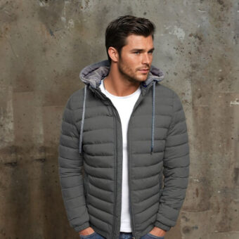 Uomo dai capelli scuri e dalla barba incolta in piedi davanti a un muro grigio scuro, indossa una maglietta bianca, jeans blu e un piumino grigio corto e sottile con cappuccio e coulisse. Le mani sono nelle tasche dei pantaloni.