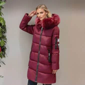 Donna bionda di profilo accanto a una parete marrone chiaro che indossa un caldo piumino bordeaux di media lunghezza con pelliccia abbinata sul cappuccio e tasche laterali. Tiene la mano destra sulla tempia. A sinistra si vede una pianta verde.