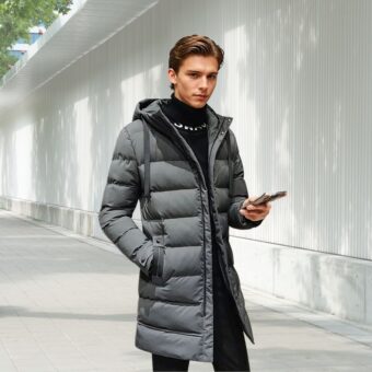 Piumino da uomo di media lunghezza di colore grigio, indossato da un giovane con i capelli castani che tiene il telefono nella mano sinistra. È in piedi per strada.