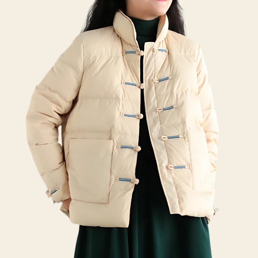 Elegante piumino da donna Tang in beige con sofisticate chiusure ad alamari, ampie tasche e collo alto. Una donna lo indossa sopra un abito nero. Le mani sono dietro la schiena.