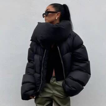 Donna che indossa un piumino nero corto con collo arrotolato oversize, chiuso da una zip argentata, maniche lunghe e larghe, in posa frontale con pantaloni neri aderenti.