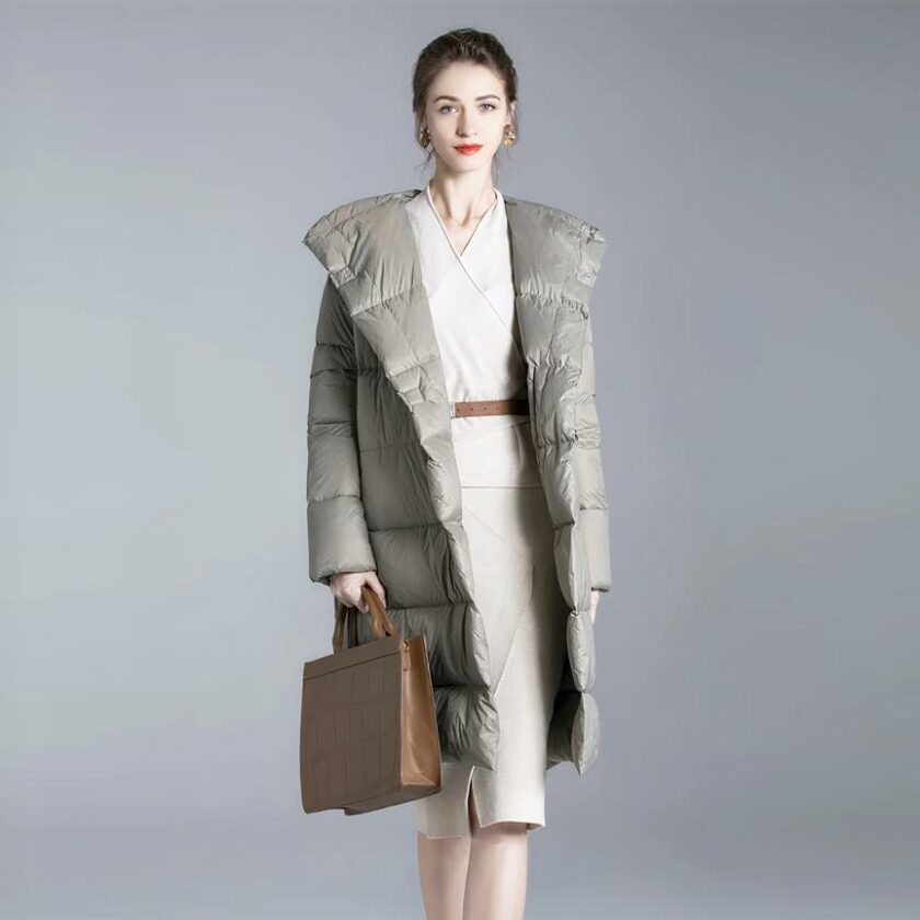 Su uno sfondo grigio, una donna con capelli castani legati all'indietro e orecchini eleganti indossa un piumino grigio lungo e largo, con una borsa marrone nella mano destra e un vestito beige lungo fino al ginocchio.