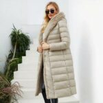Donna che indossa un lungo piumino beige con collo di pelliccia beige, chiuso da una cintura. Ha lunghi capelli sciolti e occhiali da sole, con una borsa nera in mano e scale bianche sullo sfondo.